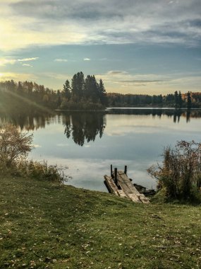 Yıpranmış ahşap iskele, sonbahar ağaçlarını yansıtan durgun bir göle doğru uzanır. Sıcak akşam ışığı sonbahar renklerini vurguluyor ve huzurlu bir deniz manzarası yaratıyor..