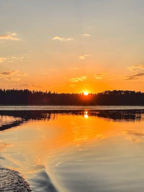 Güneş sakin bir göl üzerinde batar, gökyüzünde ve suda yoğun bir turuncu parıltı bırakır. Yansıma çarpıcı bir ayna etkisi yaratıyor, yaz akşamının güzelliğini vurguluyor..