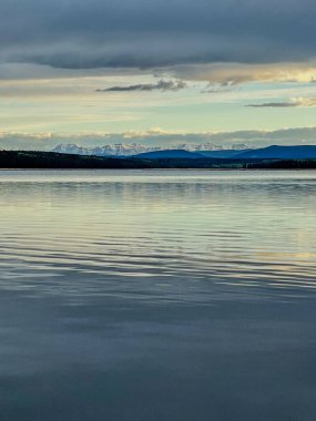 Gün batımında uzak dağları ve bulutları yansıtan sakin bir göl. Yumuşak ışık ve huzur dolu bir çevre. Sahnenin doğal güzelliği dağ yansımasıyla vurgulanır..
