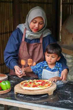 Asyalı Müslüman anne ve küçük oğlu, mutfakta birlikte yemek pişirirken masanın üstünde öksürüyorlar, tatlı küçük kız ve İslami anne evde eğleniyor, ev yapımı hamur işlerinin keyfini çıkarıyorlar.