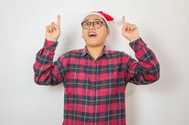 Kırmızı ekose gömlek giyen yakışıklı genç Asyalı adam ve beyaz arka planda izole edilmiş çalışma alanında işaret parmağı gösteren Noel Baba şapkası. Mutlu yıllar, tatil kavramını kutluyoruz..