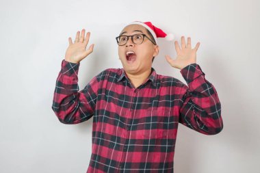 Kırmızı Noel Baba şapkası takan Asyalı adam şaşkınlığını gözler ve ağzı açık bir şekilde ifade ediyor ve ellerini şaşkına çeviriyor. Açık mavi arka planda duruyor, neşeli bir tatil görüntüsü veriyor..