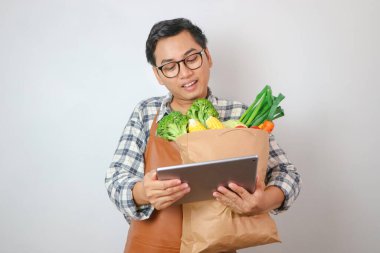 Eğlenceli Asyalı genç adam paket servis için kahverengi el çantası giyiyor. Yemek ürünleriyle maket maskesini taklit ediyor. Sade mavi arka planda izole edilmiş dijital tablet bilgisayar kullanıyor. Dükkan ya da restorandan teslimat servisi