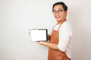 Beyaz kollu gömlek ve kahverengi önlük giyen heyecanlı Asyalı genç adam gülümseyen yüz ifadesiyle dikey beyaz ekran tableti gösteriyor. Beyaz arkaplan üzerinde izole edilmiş.