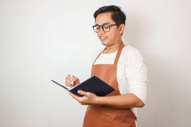 Mavi önlüklü Asyalı barista adam sipariş alıyor, gülümsüyor ve menü listesine yazıyor beyaz arka planda izole edilmiş görüntüler.