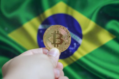 Elinde altın sarısı bir kripto para olan bir adamın eli. Elinde Brezilya bayrağının arka planında bulunan bir avuç bozuk para..