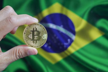 Elinde altın sarısı bir kripto para olan bir adamın eli. Elinde Brezilya bayrağının arka planında bulunan bir avuç bozuk para..