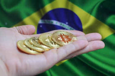 Elinde altın sarısı bir kripto para olan bir adamın eli. Elinde Brezilya bayrağının arka planında bulunan bir avuç bozuk para..
