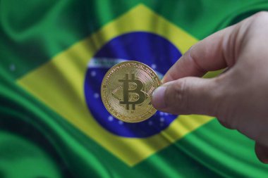Elinde altın sarısı bir kripto para olan bir adamın eli. Elinde Brezilya bayrağının arka planında bulunan bir avuç bozuk para..