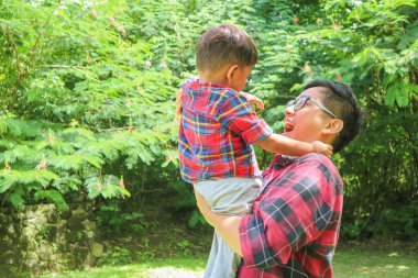 Neşeli ve mutlu Asyalı baba küçük oğluyla oturma odasında oynuyor, sırtına yaslanıyor, doğa parkında birlikte eğleniyor..