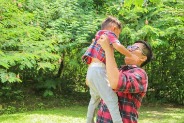Neşeli ve mutlu Asyalı baba küçük oğluyla oturma odasında oynuyor, sırtına yaslanıyor, doğa parkında birlikte eğleniyor..