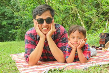 Asya aile pikniği. Baba ve oğlu, güneşli bir havada yeşil bir bahçede piknik yaparken pizza yiyorlar. Ön görünüm