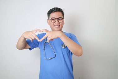 Mavi üniforma ve steteskop giyen gülümseyen profesyonel Asyalı erkek doktor ya da hemşire sarı arka planda izole edilmiş Koreli bir kalp hareketi gösteriyor. Sağlık sigortası konsepti