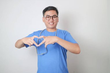 Mavi üniforma ve steteskop giyen gülümseyen profesyonel Asyalı doktor ya da hemşire beyaz arka planda elleri izole kalp şeklini gösteriyor. Sağlık sigortası konsepti