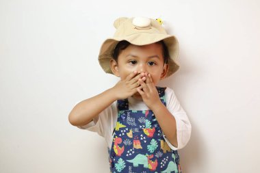 Beyaz elbiseli küçük Asyalı çocuk elini beyaz arka plana koyup ağzını kapatıyor. Koreli çocuk sessiz kalıyor ve arka planda saklanıyor.