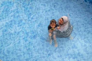 Mayo giymiş Asyalı bir çocuk annesiyle birlikte şehir binalarının arka planında yüzme havuzunda heyecanlanıyor.