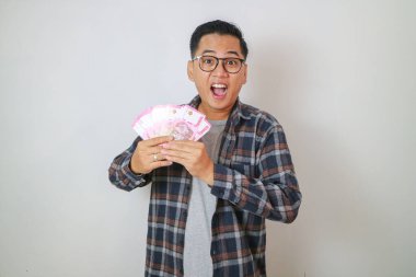 Heyecanlı genç yakışıklı Asyalı adam elinde bir deste para tutuyor. Sarı beyaz arka planda izole edilmiş.