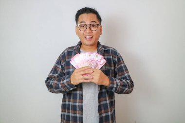 Mor arka planda izole edilmiş rupiah banknotlarla nakit para yelpazesi tutan beyaz tişörtlü mutlu genç Asyalı adam. insanlar yaşam tarzı konsepti