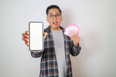 Ekose gömlekli gülümseyen genç Asyalı adam cep telefonunun boş ekranını gösteriyor ve elinde para banknotları tutuyordu, beyaz arka planda izole edilmiş kolay bir kredi aldı.