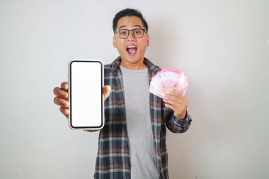 Mutlu Asyalı adam elinde Uang Endonezya parası tutuyor ve telefonu boş ekranla gösteriyor.