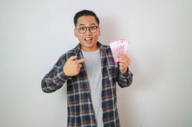 Neşeli yakışıklı Asyalı genç adam, beyaz arka planda izole edilmiş paraları işaret ediyor.