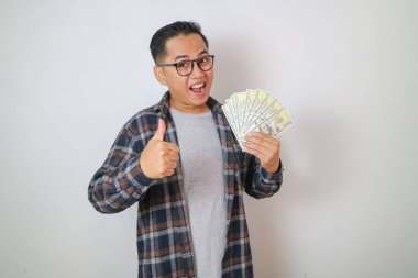 Heyecanlı gülümseyen ve gözlüklü Asyalı adam, baş parmağını sallıyor ve para sallıyor, maaş alıyor, serbest çalışıyor, iyi ödeme yapıyor, elinde nakit para tutuyor ve tatmin olmuş görünüyor.