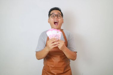 Kahverengi önlüklü, kahve dükkanında çalışan gülümseyen Asyalı genç barmen eleman, sarı arka planda izole edilmiş rupiah nakit para tutuyor. Küçük işletme başlangıç konsepti