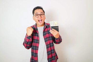 Cüzdanında para tutan ve yumruğunu kaldıran heyecanlı Asyalı genç adam beyaz arka planda izole edilmiş. Kâr ve zenginlik kavramı