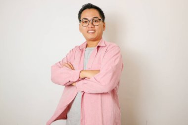 Pembe tişörtlü kendine güvenen genç Asyalı adam, çapraz kollarla poz veriyor ve beyaz arka planda izole edilmiş kameraya bakıyor..