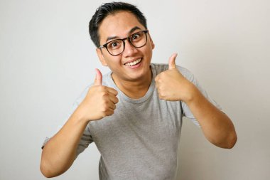 Kamerada Neşeli Asyalı Adam 'ın Başparmaklarını Göstermesi' nin yukarısında, Beyaz Stüdyo Arkaplanı 'nın önünde dikilirken, Mutlu Milenyum Erkek Hareketi Onaylama İşareti