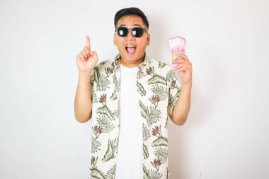 Asyalı genç adam, günlük plaj tişörtü giyip izole edilmiş beyaz arka planda durup gülümserken elinde kağıt para tutmaktan vazgeçiyor.