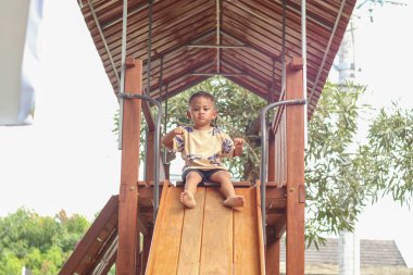 Asyalı genç bir çocuk oyun parkında ahşap sürgüde oynuyor. Mutluluktan gülümsüyor ve kollarını uçak kanadı gibi açıyor..