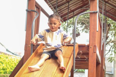 Asyalı genç bir çocuk oyun parkında ahşap sürgüde oynuyor. Mutluluktan gülümsüyor ve kollarını uçak kanadı gibi açıyor..