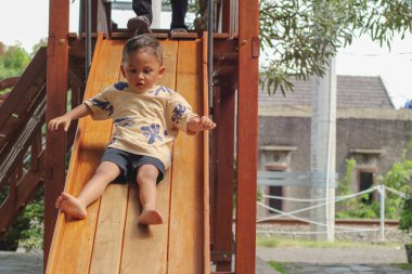 Küçük okul çocuğu, yaz mevsiminin güneşli bir gününde anaokulundaki oyun parkında tek başına kayıyor. Küçük tatlı bir çocuk, tek başına oyun sahasında kayıyor.