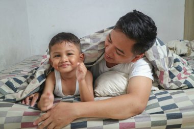 Yakışıklı, Asyalı bir baba, güzel bir Asyalı çocuğu kucaklıyor ve kucaklıyor. Evde rahat bir yatakta iyi bir gece uykusu için hazırlanıyor.