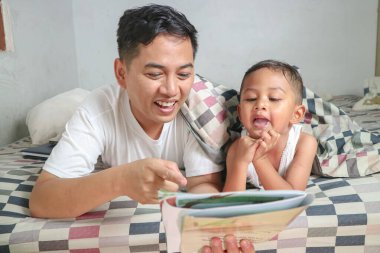 Gülümseyen baba ve şirin Asyalı küçük bebek 41 yaşında bebek masal kitabı okuyor, yatakta oturuyor, baba ve oğul birlikte vakit geçiriyor, ebeveynler çocuk konseptine kitap okuyor.