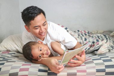 Asyalı adam ve küçük oğlu yatakta kitap okuyorlar.