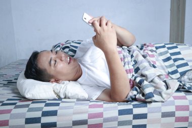 Yatakta uzanmış genç bir adam, akıllı telefonuna düşünceli bir ifadeyle bakıyor. Rahat, rahat yatak odası.