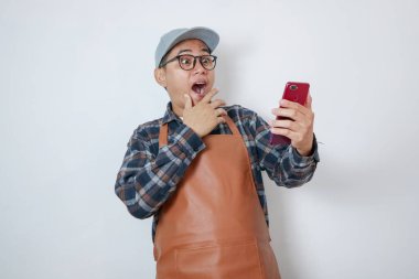 Kahve dükkanında önlük giyen genç adam cep telefonuyla sohbet ediyor ve beyaz arka planda izole bir şekilde gülümsüyor.