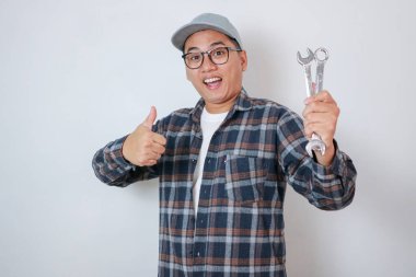 Tamirci bir ingiliz anahtarı tutarken baş parmağını kaldırıp stüdyoda duruyor, beyaz arka planda izole halde.