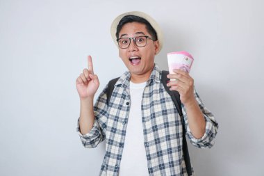 Genç Asyalı gezgin elinde kağıt para tutarken parmağını kaldırıp gülümsüyor.