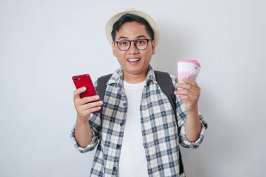 Sıradan giysiler içinde gülümseyen Asyalı genç adam portresi cep telefonuna bakıyor, beyaz arka planda izole edilmiş para tutuyor.
