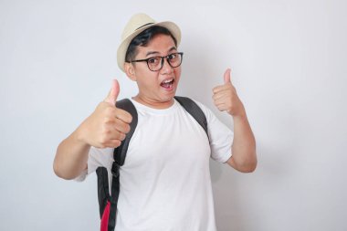 Sırt çantalı mutlu, genç Asyalı turist beyaz arka planda baş parmağını kaldırıyor.