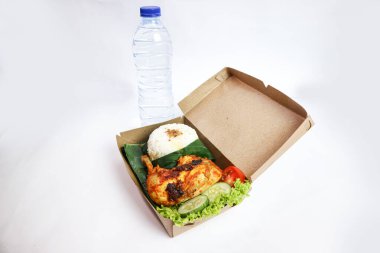 Nasi Kotak ayam bakar (kızarmış tavuk), bir tepecik beyaz pirinç ve salatalık, domates ve maden suyu gibi taze sebzelerle servis edilir.