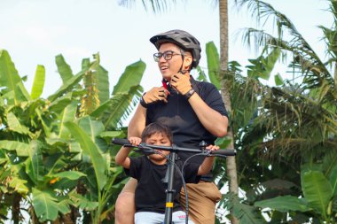 Asyalı baba ve oğul bisiklete binmeden önce kafalarına kask taktılar, oğlu ile Endonezyalı yetişkin yaz aylarında parkta birlikte aktiviteler yaptılar, baba oğul ilişkisi.