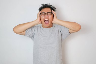 Gri t-shirt giyen, baş şakaklarına dokunan, sorunları çözmekte zorluk çeken genç Asyalı bir adam, stüdyo beyaz arka planında baş ağrısı ve panik yaşıyor..
