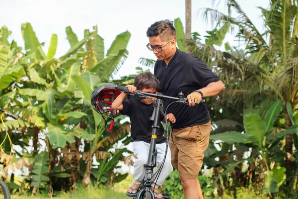 Asyalı baba oğluna bisiklet sürmeyi öğretiyor, Koreli son sınıf öğrencisi çocuğa yardım ediyor, kasklı çocuk yazın babasıyla birlikte parkta dinleniyor.