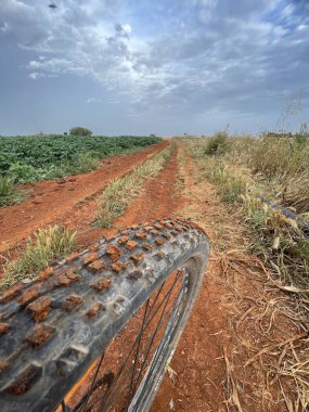 Avgorou Countryside Path A muddy bike tire rests in the foreground of a dirt path through Avgorous red-earth farmland. The surrounding fields and cloudy skies add to the sense of outdoor adventure and exploration. clipart