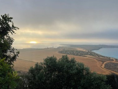 A breathtaking sunrise at Cape Greco with golden hues reflecting on the Mediterranean. Morning mist blankets the peninsula, creating a serene coastal scene. clipart