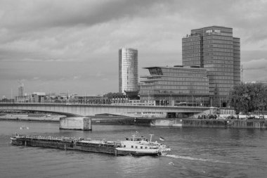 Cologne, Germany - October 27, 2023: A cargo ship cruises down the river, with modern high-rise buildings and a sleek bridge framing the scene. The calm water reflects the architecture, while the cloudy sky gives a cool and industrial vibe. clipart